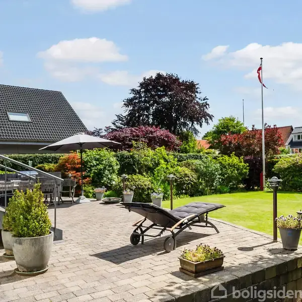 Terrasse med en sort liggestol og parasol på flisebelagt område. Omgivet af grøn have, farverige buske og flagstang. Trapper fører op til huset med et mørkt tag.
