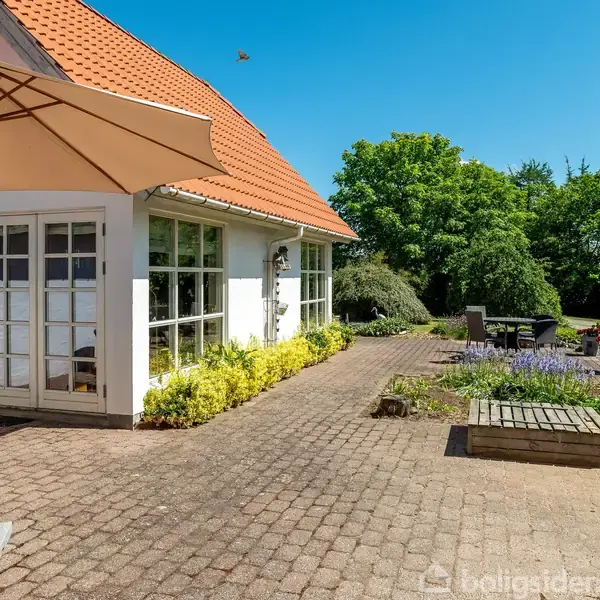 En terrasse med parasol og fliser støder op til et hus med rødt tag. Blomsterbede omgiver området, og store træer ses i baggrunden under en blå himmel.