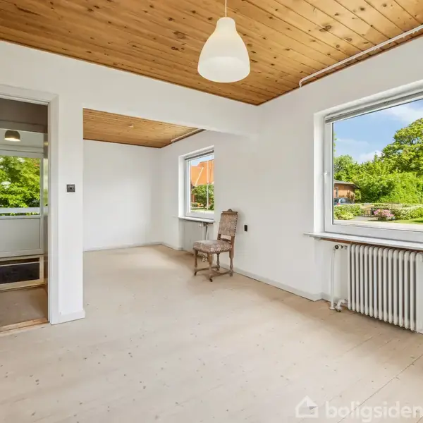 Et lyst rum med trægulv og træloft. En stol står ved et af de store vinduer, der viser en grøn have udenfor. Lampe hænger fra loftet.