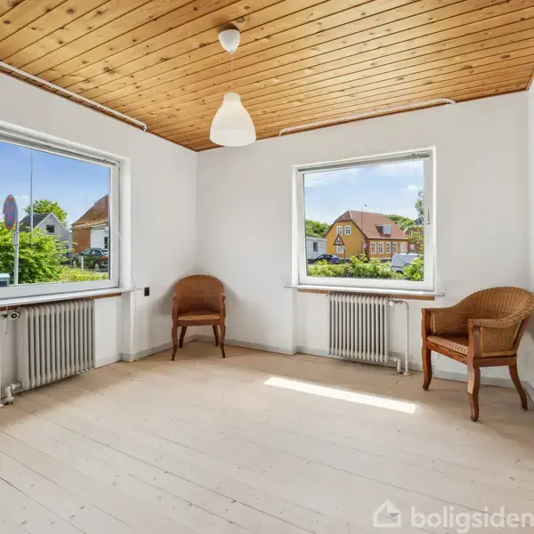 Et tomt rum med to fletstole under to store vinduer, der slipper lys ind. Trægulv og træloft med en enkelt lampe. Udsigt til huse og grønne områder udenfor.