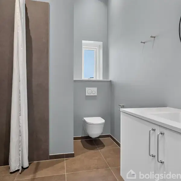 Badeværelse med en hvid vask på et skab, et væghængt toilet, og brusekabine med forhæng. Rummet har lys grå vægge, et spejl over vasken, og et lille vindue.