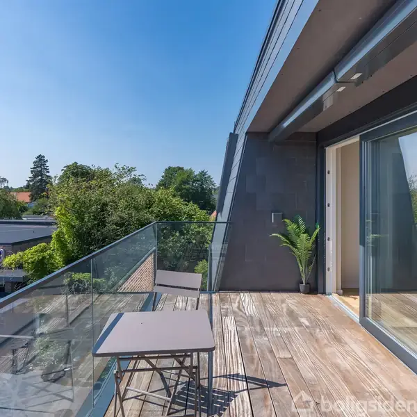 En balkon med glasrækværk rummer et bord og to stole. Den åbner til et moderne hjem med store vinduer. I baggrunden ses træer og butikskiltet "REMA 1000".