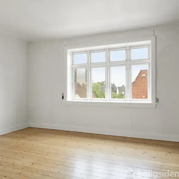 Et lyst rum med trægulv og en stor vinduesramme, der slipper naturligt lys ind. Man ser ud mod hustage og skyfri himmel. Rummet har hvide vægge og en grå accentvæg.
