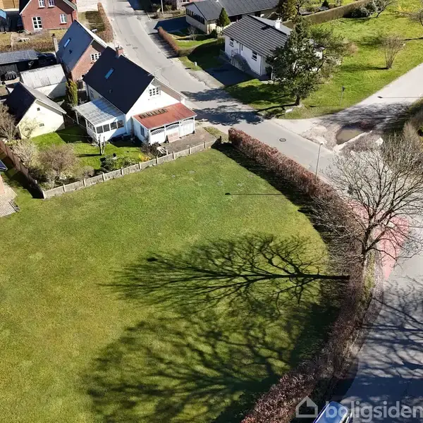 Græsplæne ligger ved vejkryds, omgivet af huse og træer, med skygge af stort træ. Beboelsesområdet har velordnede haver og flere bygninger i baggrunden.