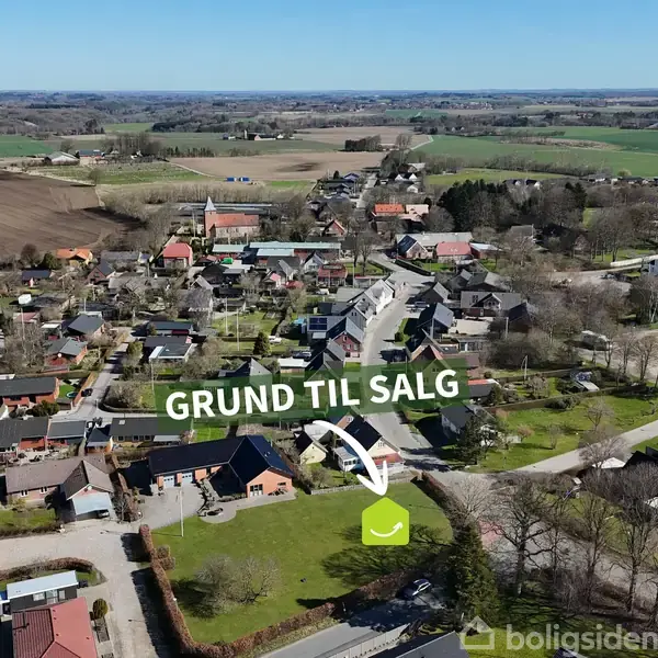 Grøn markør peger på en byggegrund, der er til salg ("GRUND TIL SALG") i en landsby omgivet af huse og åbne marker.