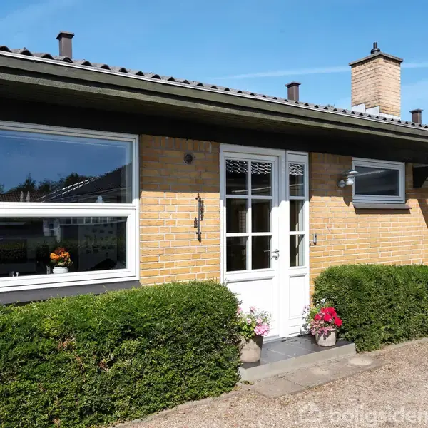 En gul murstensbygning med hvide vinduer og dør, dekoreret med blomsterpotter. Foran er der en lav hæk og en grusbelagt indkørsel under en klar blå himmel.