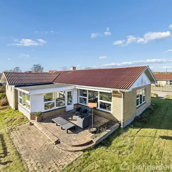 Et enfamiliehus med rødt tegltag står i en grøn have. Huset har store vinduer og en terrasse med møbler, beliggende i et villakvarter under en blå, skyet himmel.