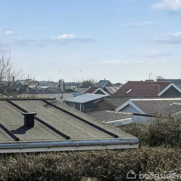 Flade tage med skorstene strækker sig over et boligområde med buske i forgrunden og en klar blå himmel i baggrunden.