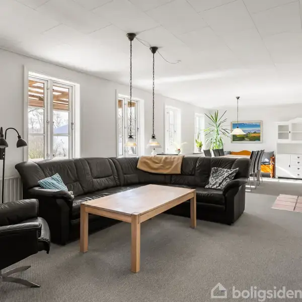 Sort læderhjørnesofa står i stue med hvide vægge, omkring et træbord. Loftslamper hænger over sofaen. I baggrunden ses vinduer, planter og en spiseplads med maleri på væggen.