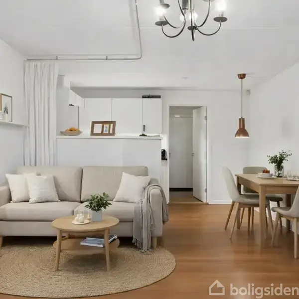 Afdæmpet opholdsstue med grå sofa ved et rundt sofabord, placeret på et tæppe. Til venstre står en gulvlampe, bagved et lille, hvidt køkken. Spiseområde med bord og stole til højre. Øverst står teksten "Illustration".