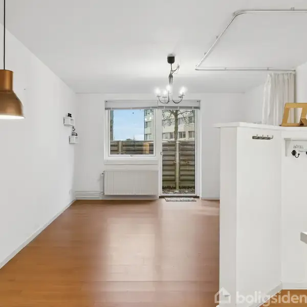 Et tomt rum med trægulv og hvide vægge. En kobberlampe hænger fra loftet. Der er også et lille vindue og en dør foran, der fører ud til en terrasse.