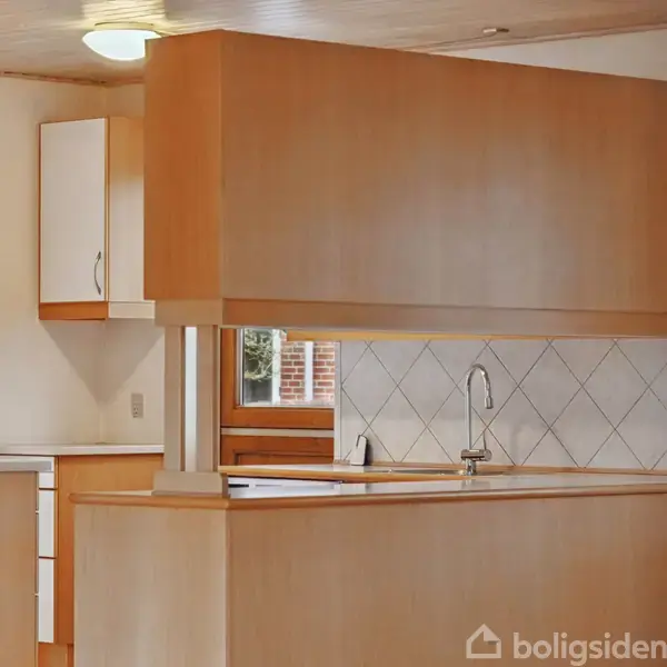 Køkken med træskabe og hvidt flisebaggrund, indeholder en vask med vandhane. Set fra en indgang viser det en lys, ryddelig atmosfære med naturligt lys fra et vindue.