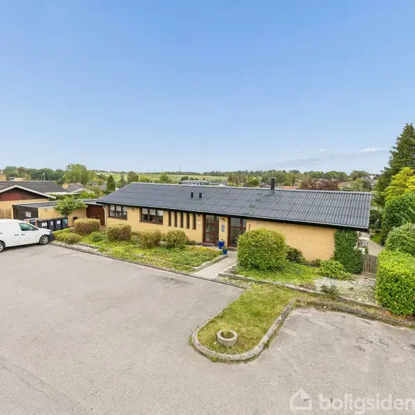 Et enfamilieshus med fladt tag står ved siden af en parkeret hvid bil på en rolig gade. Huset er omgivet af grønne buske og træer i en landlig område.