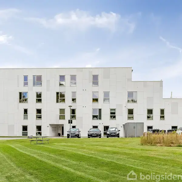Hvid bygning står stille, omgivet af grønne græsplæner med parkerede biler foran. Bag ved bygningen er der træer og buske, der giver en naturlig baggrund.