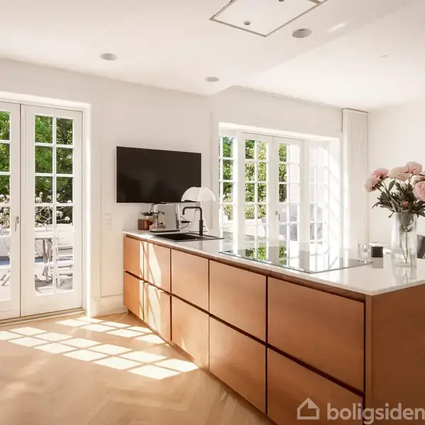 Køkkenø med bruntræsbasis og hvid bordplade står i sollys, omgivet af dobbeltdøre og vinduer. En vase med blomster og en vægmonteret skærm er opstillet på øen.