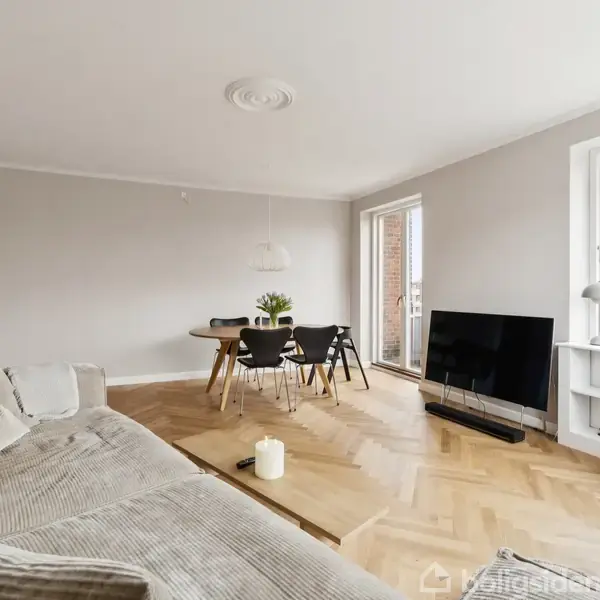 En lys stue med en stor sofa, et fladskærms-TV på gulvet, og et spisebord med fire stole. Rummet har parketgulv og store vinduer mod en altan.ingen tekst.