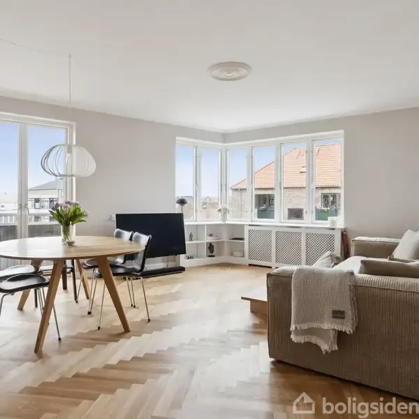 Stue med en sofa og et rundt spisebord med stole. Et stort vindue giver udsigt til bygninger udenfor. Lyse parketgulve og minimalistisk indretning skaber et åbent miljø.