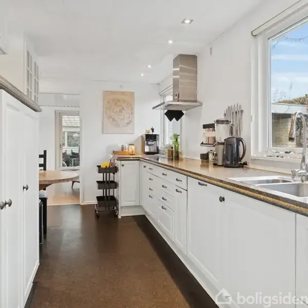 Hvidt køkken med apparater som elkedel og kaffemaskine står på en lang bordplade. Store vinduer lader naturligt lys ind. I baggrunden ses spiseområde.