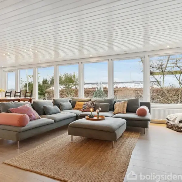 En stor grå sofa med farverige puder står i en lys stue med trægulv. Store vinduer giver udsigt til en have, og der er et spisebord i baggrunden.