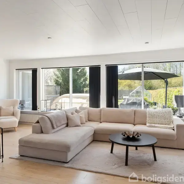 Stor, lys stue med en L-formet beige sofa, der omgiver et rundt sofabord. Store vinduespartier giver udsigt til en terrasse med parasol. Lyse trægulve og moderne indretning.