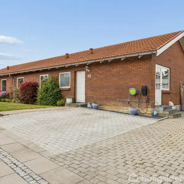 Rød murstensbungalow med hvid hoveddør nummer 45; flisebelagt indkørsel, ledninger på væggen; omgivet af græsplæne, buske, hegn og skur. Klar blå himmel.