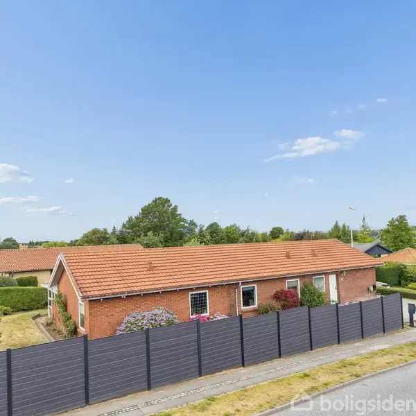 Et rødt murstenshus med rødt tegltag er omgivet af en sort hegn. Huset ligger i et beboelsesområde med grønne træer og blå himmel i baggrunden.