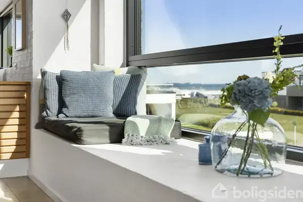 Vindueskarmen er dekoreret med blå puder og tæppe, og en glasvase med blomster. Sollys strømmer ind, mens en udsigt over græsarealer og bygninger kan ses udenfor.
