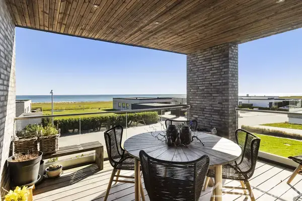 Rundt bord med sorte stole står på en træterrasse under et tag. I baggrunden ses moderne huse, grønne områder og havet under en klar, blå himmel. Potteplanter er på terrassen.