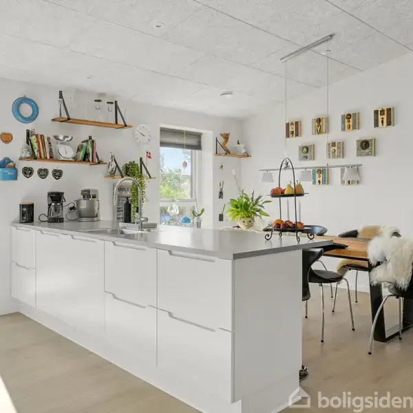 Et moderne køkken med en hvid køkkenø står i centrum, omgivet af åbne hylder og dekorative elementer. Store vinduer lader lys ind, med udsigt til en terrasse.