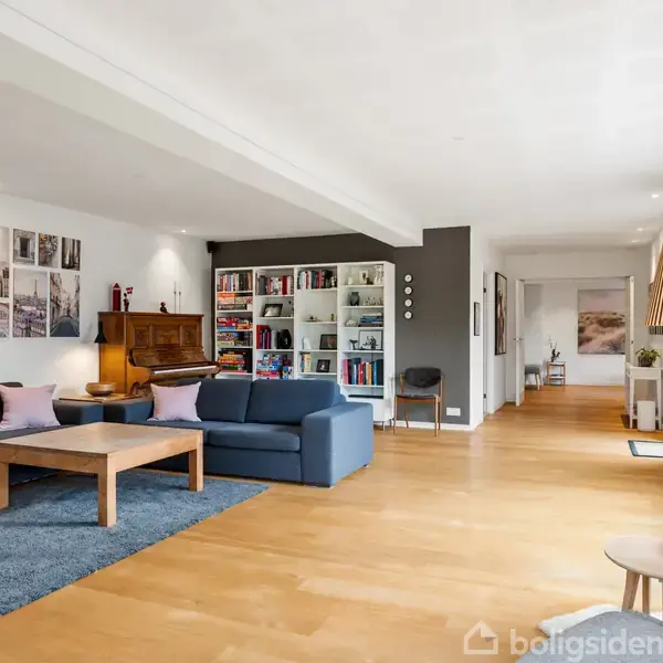 Stue med blå sofa, træbord og tæppe. Reoler fyldt med bøger i baggrunden. Trægulv og stort vindue til højre med lamper og blomster. Indretning er moderne og indbydende.