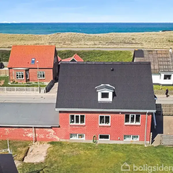 Rødt murstenshus med sort tag står i sollys, omgivet af græsplæne og indhegnet have. Havet og klitterne kan ses i baggrunden, mens flere huse står langs vejen.