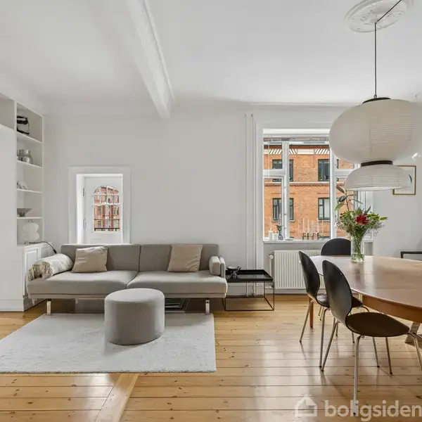 Stue med grå sofa og puf på tæppe, spisestue med stort træbord og stole. Store vinduer giver naturligt lys, hvide vægge, og trægulv. Blomster og lampe tilføjer dekoration.