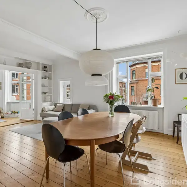 Rundt bord med stole står i lyse stue. Stor lampe hænger over bordet. Sofa ved hylder i baggrunden. Vindue viser udsigt over bygninger. Blomster og dekorationer pynter rummet.