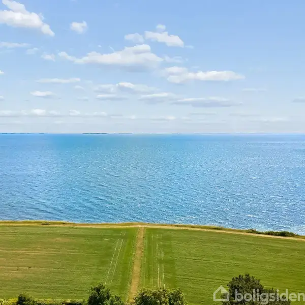 Et rødt pil peger mod et hus i en beboelsesområde nær kysten. Baggrunden viser en bred udsigt over havet og kystlinjen med grønne marker.