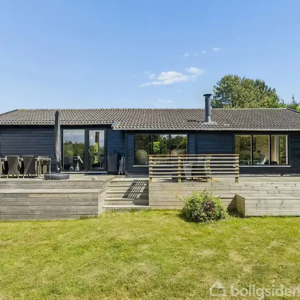 En moderne træhus med terrasse omgivet af græsplæne og træer. Huset har store vinduer og en skorsten, mens møbler står på terrassen. Klatreskubber afslutter terrenget mod haven.