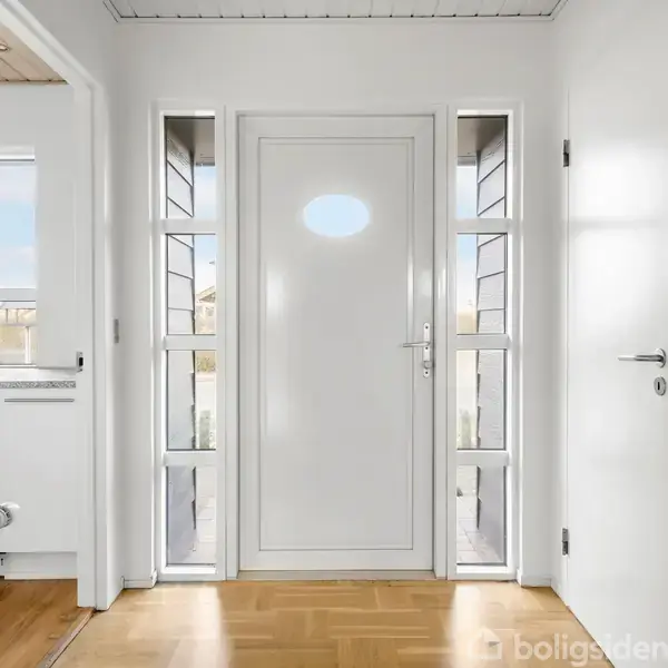 Dør står mellem to vinduer med rektangulært mønster i en entré. Der er trægulv, hvide vægge, og en radiator til venstre. En smal udsigt til udendørs ses gennem vinduerne.