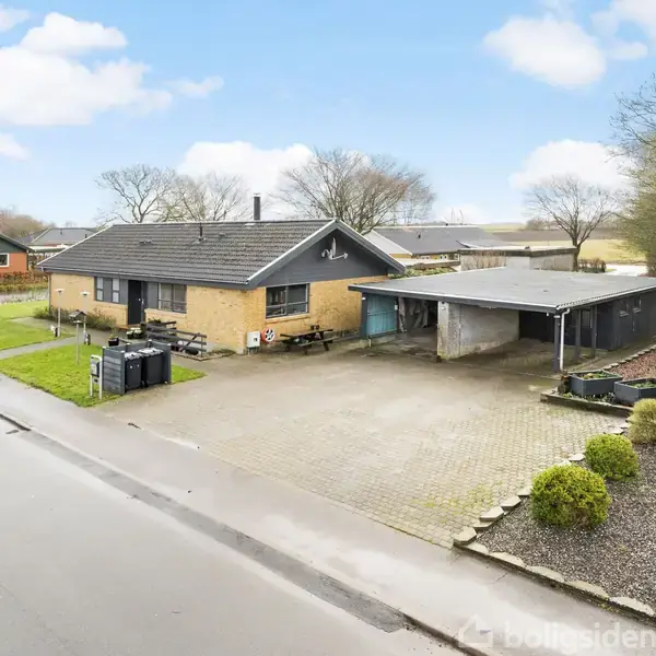 Et enfamiliehus af mursten står stille ved en stille vej. Omkring huset ses en indkørsel med carport og en græsplæne suppleret af buske og træer.