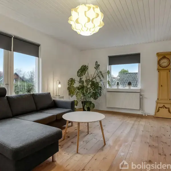 Sort sofa står i stue med lyse træplanker og loft. Rundt hvidt sofabord foran, stor lampe hænger ovenover. En gulvplante ved vindue og gammelt ur i hjørnet.