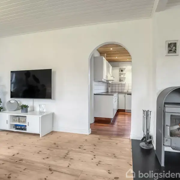 En moderne stue med væghængt TV og et bord, imens en tændt brændeovn står ved siden af. Gennem en bue ses et lyst køkken med træog gulv.