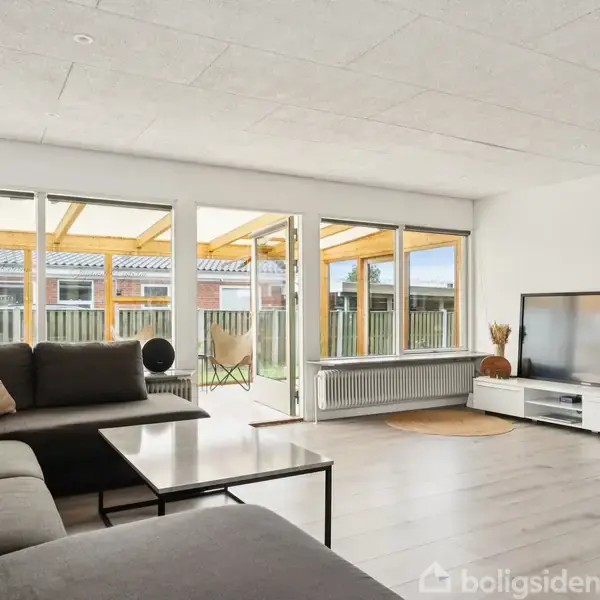 En stue med en stor grå sofa og et sofabord. TV står på et lavt skab. Rummet er lyst med store vinduer mod en terrasse.