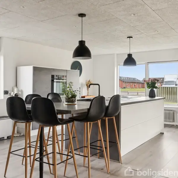 Et moderne køkken med en ø, omgivet af høje sorte stole. Køkkenøen har en hvid top med planter og lamper hænger over. Store vinduer giver udsigt til en have.