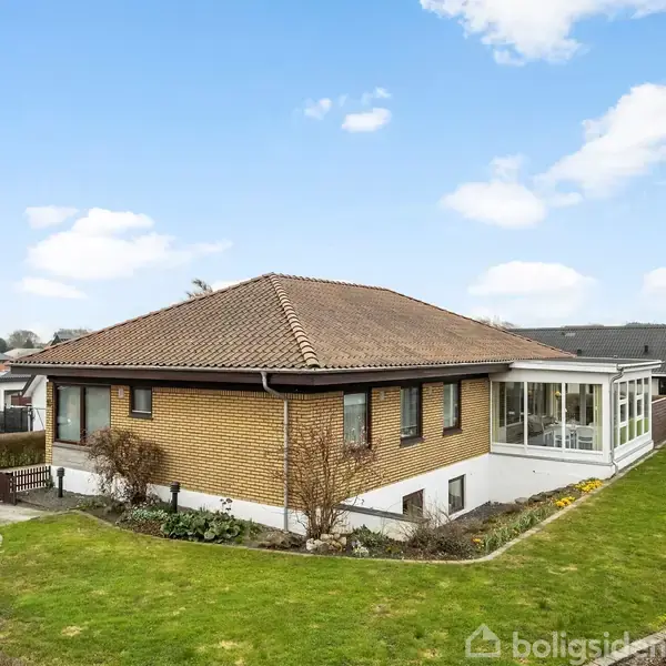 Huset er en etplans villa med brun murstensfacade og rødt tegltag, omgivet af en velplejet græsplæne og en hæk. Ved siden af huset står en flagstang.