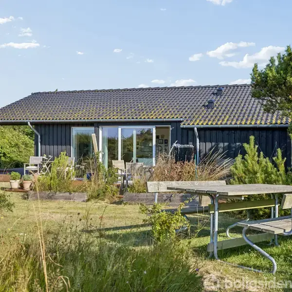 En sortmalet bungalow har store vinduer og terrasse med havemøbler, omgivet af grønt græs og buske. En bænk med bord står i forgrunden under en klar blå himmel.