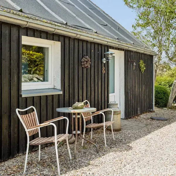 En lille gårdsplads har et sort træhus med et hvidt vindue og en dør. To stole og et bord står udenfor. En lille sti fører langs huset.