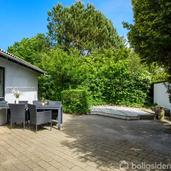 Terrasse med bord og fire stole, omgivet af frodig have og træer. Til venstre er en hvid bygning med et åbent vindue, mens en platform ses i baggrunden.