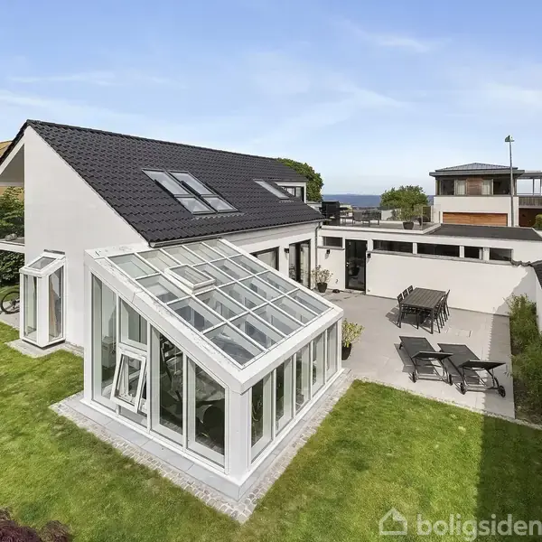 Et moderne hus med et glastilbygning ligger i en grøn have. En terrasse med havemøbler ses til højre, og der er udsigt til havet i baggrunden.