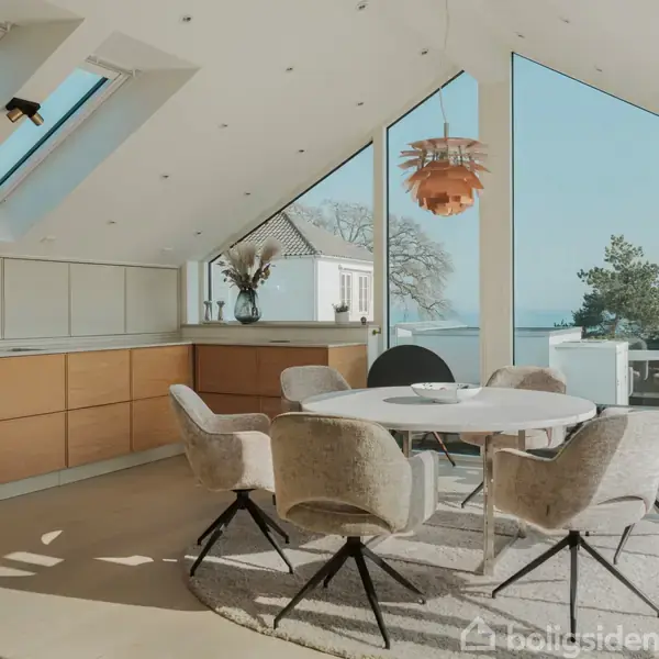 Lysegråt spisebord med seks polstrede stole står i et moderne køkken-alrum. Store vinduer og ovenlysvinduer lukker naturligt lys ind. Udsigt til træer og vand i baggrunden.