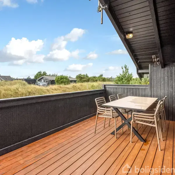 Terrasse med sortmalede trævægge og træbord i centrum, omgiven af seks stole. Udsigt over græsbevokset område med huse i baggrunden under en blå himmel med få skyer.