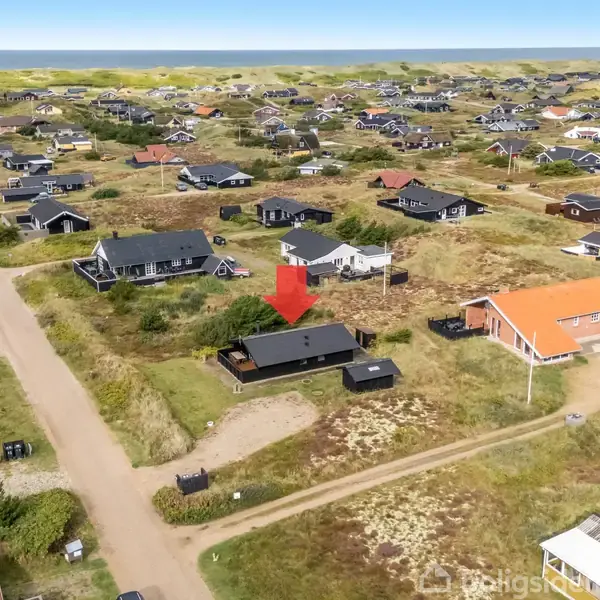 Hus markeret med rød pil er beliggende blandt flere huse i et åbent, grønt område med sandveje. Landskabet er fladt og præget af klitagtig vegetation nær kysten.