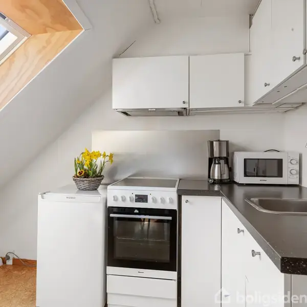 Et hvidt køkken har en ovn ved siden af et lille køleskab med en kurv med blomster ovenpå. En kaffemaskine og mikrobølgeovn står ved siden af vasken.